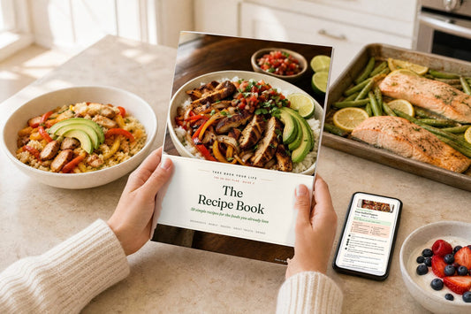 Hands holding The Recipe Book from the Take Back Your Life 28-Day Plan, surrounded by fajita bowls, sheet pan salmon and a phone showing a recipe